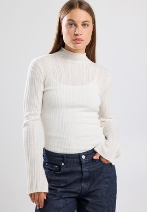 Camiseta blanca de manga larga con cuello alto y puños acampanados, combinada con jeans de rayas azul oscuro. Textura suave, diseño ajustado.