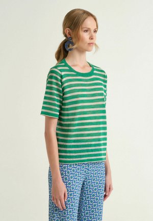 Donna con capelli biondi raccolti in una coda bassa, indossa un maglione a maniche corte a righe verdi e bianche, pantaloni blu con motivo e grandi orecchini blu.