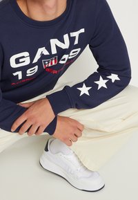 Marineblauw sweatshirt met witte grafische tekst "GANT 1999" en sterren op de mouwen; gecombineerd met crèmekleurige broek en witte sneakers.