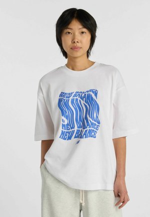 Ung person med kort sort hår bærer hvid oversized New Balance t-shirt med blåt forvrænget logo og lysegrå sweatpants, stående.
