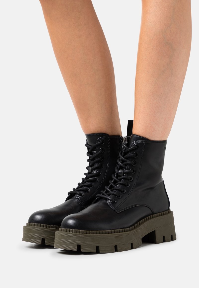 Bottes de combat en cuir noir à lacets avec semelles vertes épaisses portées sur des jambes nues sur un fond clair uni.