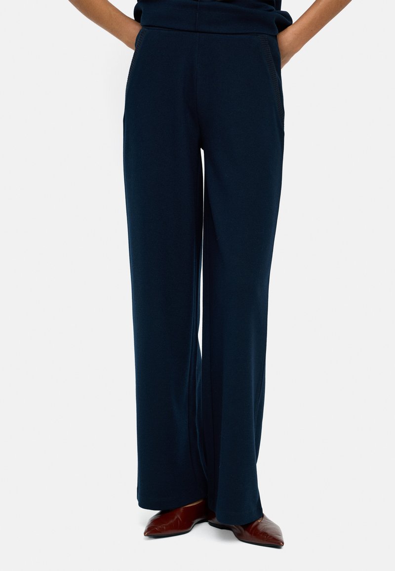 Granatowe spodnie o szerokich nogawkach z miękkiego materiału, z wysokim stanem i kieszeniami bocznymi, zestawione z brązowymi butami na szpilce.