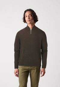 Cárdigan marrón con cremallera hecho de una tela suave, con un alto cuello y puños acanalados. Combinado con pantalones de un verde oliva claro.