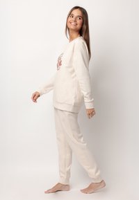 Joven sonriendo, caminando descalza en ropa de casa beige con un sudadera gráfica floral contra un fondo blanco liso.