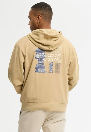 Hombre con sudadera beige con capucha con una formación rocosa ilustrada y coordenadas impresas en la espalda, de pie frente a un fondo blanco liso.
