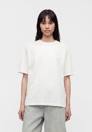 Kvinde med langt mørkt hår iført en almindelig hvid kortærmet t-shirt og lysegrå bukser, der står mod en hvid baggrund.