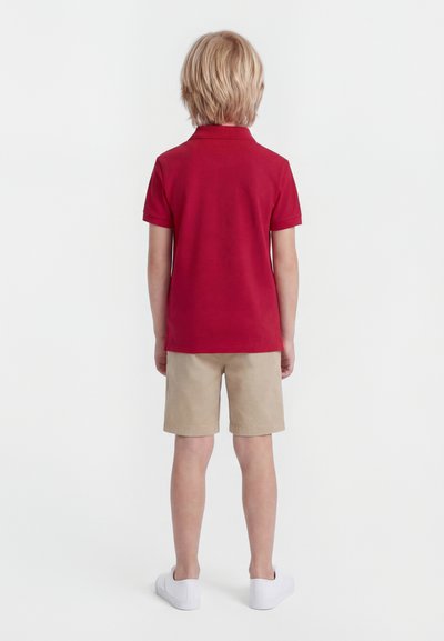 Heledate juustega laps seisab seljaga vaataja poole, seljas punane lühikeste varrukatega polosärk, beežid lühikesed püksid ja jalas valged libisevad jalanõud, taust on valge.