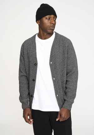 Grå ribbet cardigan med fem brune knapper, båret over en hvid t-shirt. Modellen har en sort strikket beanie på. Enkel, afslappet design.