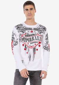 Weißes Langarm-T-Shirt mit schwarz-rot grafischen Designs, das Sterne, Totenköpfe und ein zentrales Emblem zeigt. Hergestellt aus einem glatten Stoff.