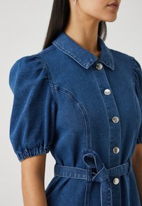 Καπιτονέ φόρεμα από τζιν με γιακά, φουσκωτά κοντά μανίκια, ασημένια κουμπιά και ζώνη στη μέση, σε σκούρο μπλε ύφασμα με λεπτή υφή.