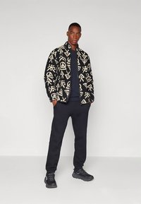 Černá prošívaná bunda s béžovými grafickými vzory, doplněná tmavou košilí a černými tepláky. Model nosí černé sportovní boty.