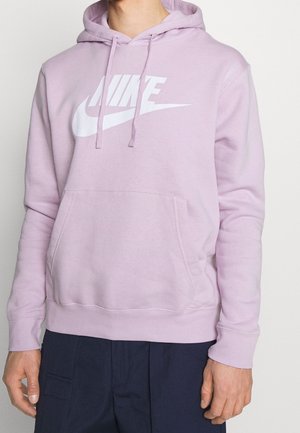 Mann trägt hellvioletten Nike-Hoodie mit Fronttasche und weißem Logo, kombiniert mit dunkelblauer Hose, steht vor weißem Hintergrund.