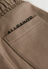 Beiger Stoff mit strukturierter Oberfläche und elastischem Bund. Verfügt über eine abgesteppten Tasche mit dem in Schwarz gedruckten Schriftzug "ALL SAINTS".