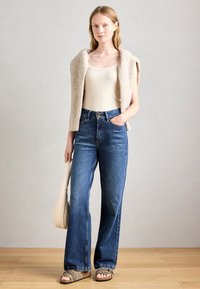 Donna che indossa un top beige, un maglione beige lavorato a maglia drappeggiato sulle spalle, jeans blu a vita alta, sandali e tiene una borsa beige.