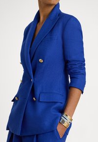 Dvojno zapetan blazer kraljevsko modre barve s teksturo ribje kosti, z zlatimi gumbi in stranskimi žepi. Na zapestju so zložene zlate zapestnice.