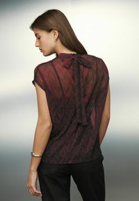 Red and black patterned blouse with short sleeves, a round neckline, and a bow detail at the back, paired with black pants.