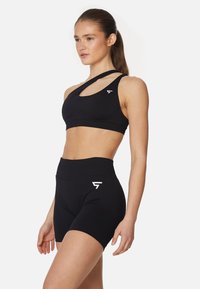 Črna športna modrček z edinstvenim križnim trakom in črne hlače z visokim pasom. Oboje je izdelano iz gladkega, raztegljivega materiala z logotipom.