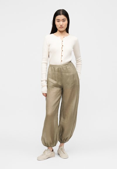 Femme debout, portant un haut blanc à manches longues boutonné, un pantalon ample vert olive avec des poignets élastiques et des baskets beiges sur un fond uni.