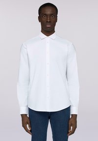 Camicia bianca a maniche lunghe con colletto a punte. Realizzata in un tessuto morbido, presenta un orlo curvo e un design semplice. Indossata con jeans scuri.