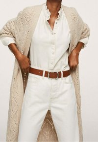 Beżowy dziergany kardigan na białej bluzce z falbanianym kołnierzykiem, zestawiony z białymi, wysokim stanem jeansami oraz brązowym skórzanym paskiem.