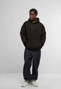 Musthave suuruses must kapuutsiga särke, millel on eesmine tasku ja iseloomulik logo. Kombineeritud laia tumeda teksapükste ja valgete tossudega. Lihtne taust.