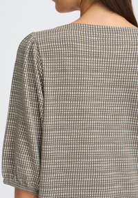 Blouse à carreaux brun et blanc avec une coupe décontractée, un col rond et des manches bouffantes. Le tissu a une texture douce.