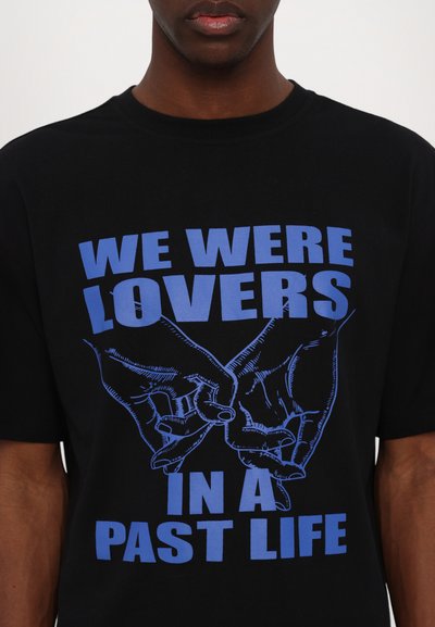 Schwarzes Baumwoll-T-Shirt mit blauer Grafikschrift: "WIR WAREN LIEBESPAARE IM FRÜHEREN LEBEN" und einer Illustration von zwei Händen, die sich miteinander verweben.