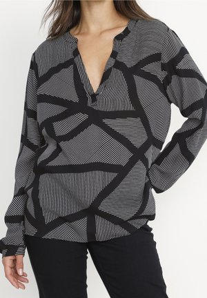 Woman wearing a black and white long-sleeve blouse with abstract geometric lines and black pants, hand on hip.