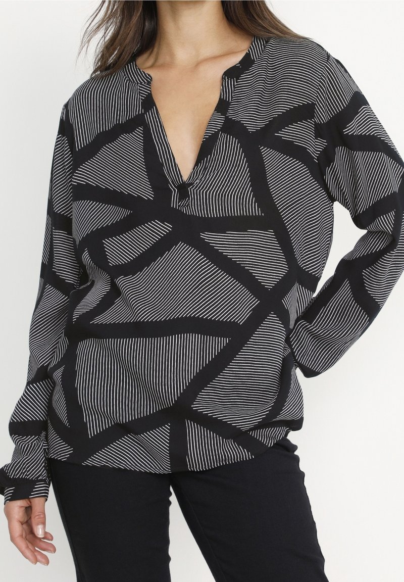 Femme portant un chemisier à manches longues noir et blanc avec des lignes géométriques abstraites et un pantalon noir, main sur la hanche.