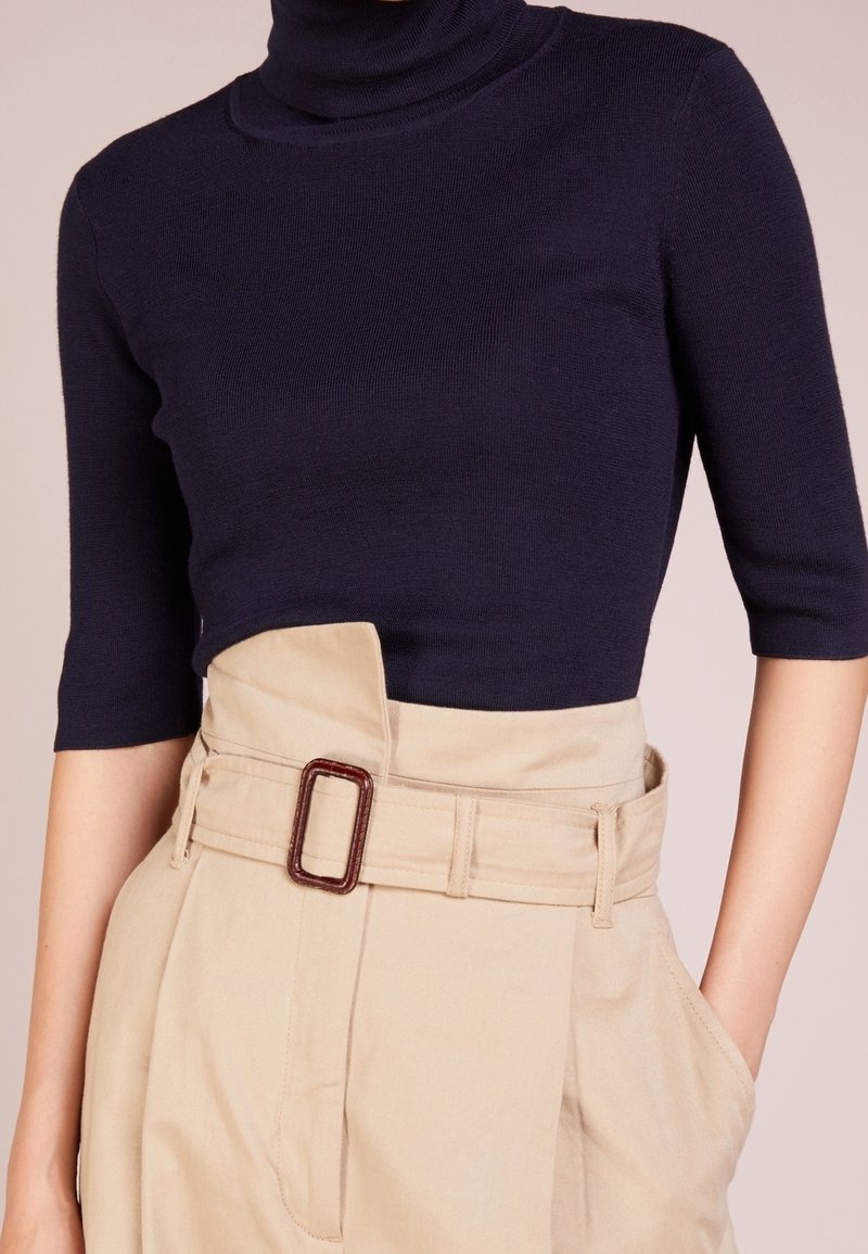 Navy-blauer Rollkragenpullover mit kurzen Ärmeln, kombiniert mit beigen hoch-taillierten Hosen, die einen breiten Gürtel mit rechteckiger Schnalle tragen.