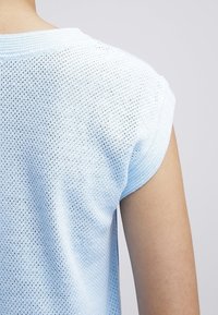 Svetle modrý pletený top s volnou, otevřenou strukturou. Má kulatý výstřih a krátké rukávy, které ukazují texturovanou látku.