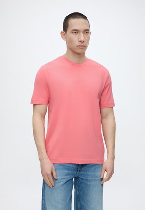 Άνδρας με κοντά μαλλιά που φοράει απλό κοραλί κοντομάνικο μπλουζάκι και ανοιχτό μπλε τζιν, στέκεται μπροστά σε λευκό φόντο.