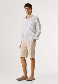 Jaunas vyras stovintis, dėvintis baltą dryžuotą ilgomis rankovėmis marškinius, smėlinės spalvos krovinines šortus ir smėlinės spalvos slip-on batus, priešais paprastą foną.