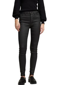 Femme portant un jean skinny noir taille haute avec une fermeture à double bouton, associé à des mocassins noirs et un haut à manches longues noir.