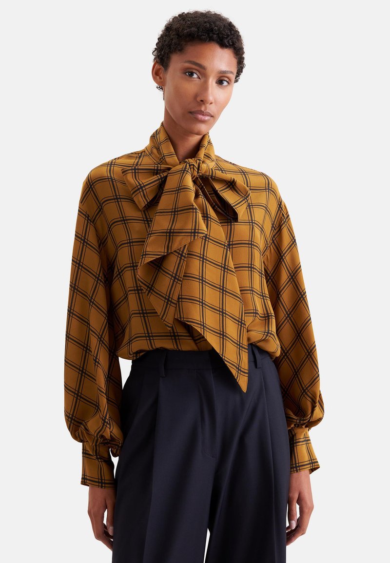 Femme portant un chemisier à carreaux jaune moutarde avec un grand nœud au col et un pantalon taille haute bleu marine sur fond uni.