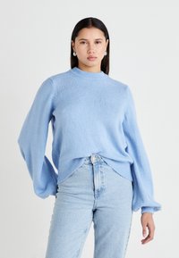 Lyseblå strikkegenser med høy hals, lange ballongstil-ermer og en avslappet passform. Kombinert med høytlivede lyseblå jeans.