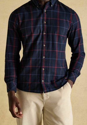 Mężczyzna ubrany w granatową, czerwoną i czarną koszulę w kratę na guziki oraz beżowe spodnie sztruksowe, stoi z jedną ręką w kieszeni.