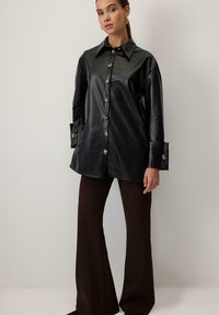 Sort faux læder skjorte med spids krave, lange ærmer, metalliske knapper og brede manchetter, parret med brune flared bukser.