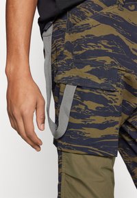 Primer plano de una mano al lado de pantalones cargo con patrón de camuflaje, que tienen grandes bolsillos y bucles de tela gris adjuntos.