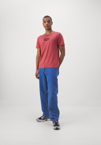 T-shirt vermelho de manga curta com o logotipo preto da Levi's na frente, combinado com jeans baggy azul claro e ténis pretos com detalhes vermelhos.