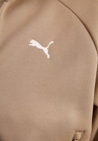 Beige athletic jacket with a textured fabric. Features a white Puma logo positioned on the chest and a zippered collar.