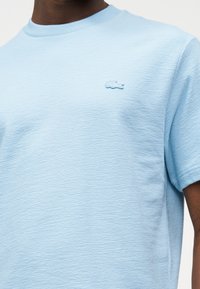 Homme portant une chemise à manches courtes texturée bleu clair avec un petit logo crocodile bleu assorti sur le côté gauche de la poitrine.