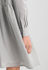 Randig skjortklänning i ljusgrått och vitt, med långa ärmar med knäppta manschetter och en utsvängd kjol, gjord av lättviktigt tyg.