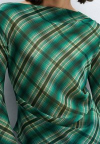 Donna che indossa una blusa lungo-sleeve a quadri verde e beige con un dettaglio di tessuto attorcigliato e drappeggiato in vita.