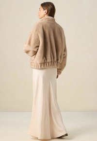 Veste beige, douce et duveteuse avec un col montant et une taille élastique, portée sur une jupe en satin fluide de couleur claire. Texture lisse avec une coupe décontractée.