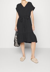 Vestido negro de longitud hasta la rodilla con mangas cortas y escote en V. Presenta una cintura ceñida con cinturón y un dobladillo con volantes. Complementado con un bolso ligero y zapatos de color claro.