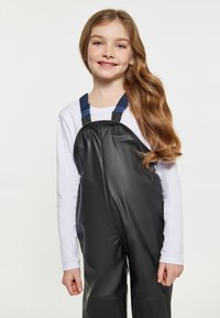 Ung flicka med långt vågigt hår, iklädd en vit långärmad skjorta och svarta vattentäta overaller med blå hängslen, leende mot kameran.