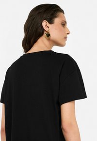 Zwarte t-shirt met een losse pasvorm, korte mouwen en een ronde hals. Gouden hartvormige oorbellen zijn zichtbaar. Glad stoftextuur.