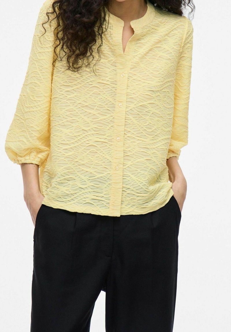 Femme portant une blouse texturée jaune clair à boutons avec des manches trois-quarts et un pantalon noir, les mains dans les poches, cheveux bouclés visibles.