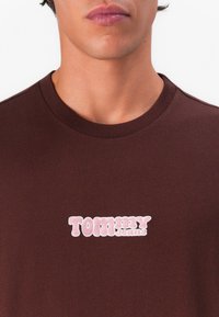 Brown cotton t-shirt featuring a pink embroidered "TOMMY JEANS" logo. Round neckline and short sleeves, with a smooth texture.
